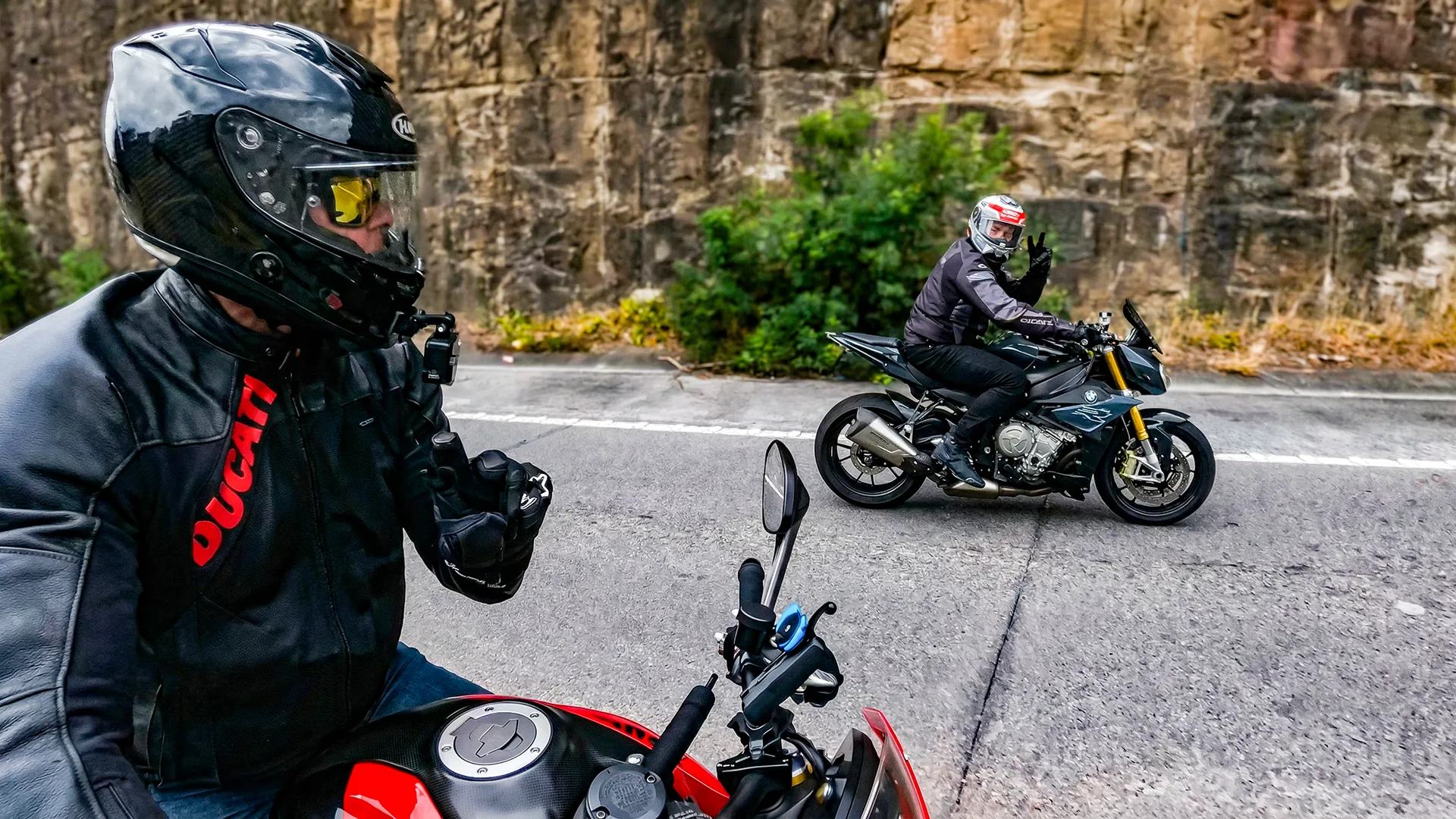 Two riders stopped on a mountain road, one signalling to the other